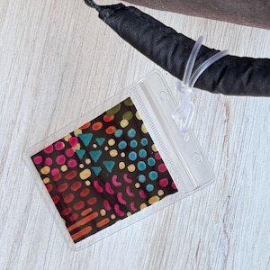 May include: A clear plastic badge holder with a colorful patterned insert. The pattern features a variety of shapes and colors, including red, yellow, blue, and green.