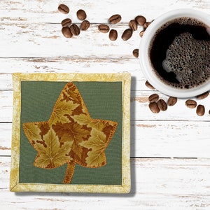 May include: A square, green fabric coaster with a leaf design in shades of brown and gold, framed with a gold border. A white coffee cup filled with dark coffee and coffee beans are on a white wooden surface.