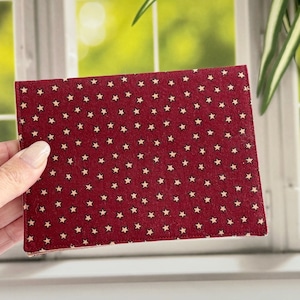 May include: A rectangular, burgundy-colored fabric item with a repeating pattern of small, cream-colored stars. The item is held in a person's hand, with a window and greenery in the background. The item appears to be a small notebook or a similar craft item.
