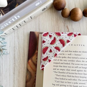 May include: A white fabric bookmark with a red watermelon pattern. The bookmark is folded in half and is placed on a book with the text "CHAPTER NINE" visible. The text "not be - well - human." is also visible on the page.