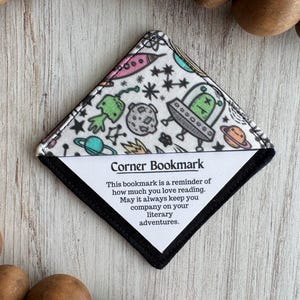 May include: A corner bookmark with a black border and a white interior. The top fabric features a colorful space-themed pattern with planets, aliens, and spaceships. Text on the bookmark reads: "Corner Bookmark...literary adventures."