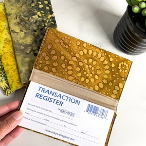 May include: A yellow and brown fabric checkbook cover with a transaction register inside. The register has a barcode and the text "TRANSACTION REGISTER" and "EASYREAD".