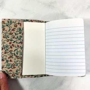 May include: A small notebook with a floral patterned fabric cover. The notebook has lined pages and is open to reveal the blank pages.
