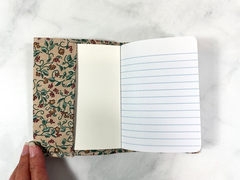 May include: A small notebook with a floral patterned fabric cover. The notebook has lined pages and is open to reveal the blank pages.