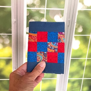 May include: A handmade, patchwork wallet with a navy blue cover and a colorful, patterned center. The center features a grid of red and blue squares, with paisley and floral designs. The wallet is held in a hand.