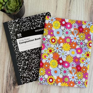 May include: Two composition books on a wooden surface. One is black and white with the text "Wide Ruled Composition Book". The other has a floral pattern with yellow, pink, orange, and blue flowers.