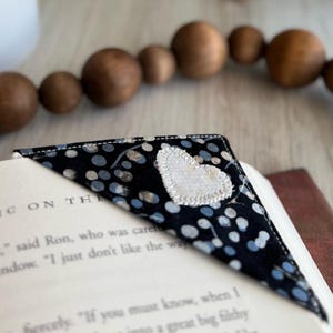May include: A handmade fabric bookmark with a black background and a pattern of blue and white dots. A white heart is sewn onto the corner. The bookmark is placed on the corner of a book with text visible.