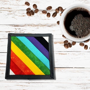 May include: A rainbow-colored fabric coaster with a black border. The coaster is on a white wooden surface with coffee beans scattered around it. A white coffee mug with black coffee is also visible in the image.