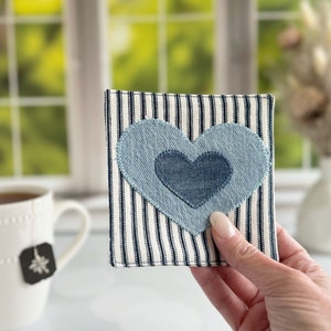 May include: A handmade coaster features a denim heart appliqué on a navy and white striped fabric. The heart is layered, with a smaller, darker blue heart inside a larger, lighter blue heart.  The coaster is square and appears to be made of a sturdy, possibly cotton, fabric. This unique coaster is perfect for adding a touch of rustic charm to any home décor.