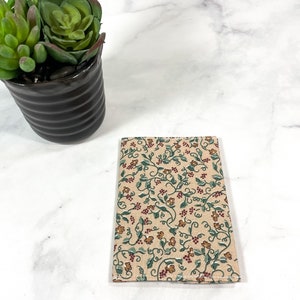 May include: A small, beige fabric notebook cover with a floral pattern of green vines, red berries, and small orange flowers.