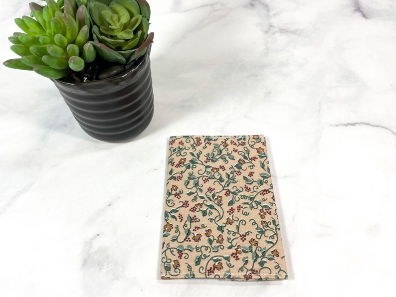 May include: A small, beige fabric notebook cover with a floral pattern of green vines, red berries, and small orange flowers.