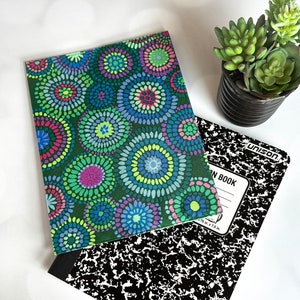May include: A composition notebook with a colorful, patterned cover featuring a design of concentric circles in shades of green, blue, purple, and pink. A black and white composition book and a small potted succulent are also present.