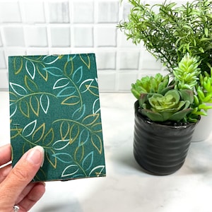 May include: A green fabric with a white and gold leaf pattern. The fabric is being held by a hand.