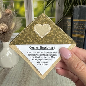 May include: A handmade corner bookmark with a heart applique. The bookmark features a green floral pattern and the text "Corner Bookmark" with an inspirational message. The bookmark is held in front of a window and books.