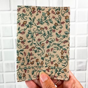 May include: A beige fabric wallet with a floral pattern featuring green vines, brown flowers, and red berries. The wallet has a stitched edge.