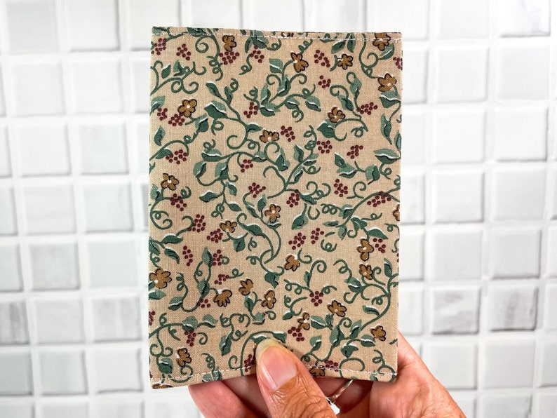 May include: A beige fabric wallet with a floral pattern featuring green vines, brown flowers, and red berries. The wallet has a stitched edge.