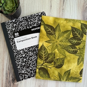 May include: A black and white composition book with the text "Wexford Wide Ruled Composition Book" and a green leaf-patterned book cover. The composition book has 80 sheets. A small potted succulent is in the background.
