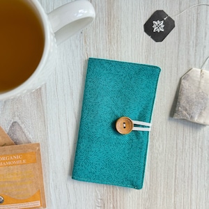 May include: A teal blue fabric phone case with a white button and a white elastic band closure. The case has a white and teal floral pattern.