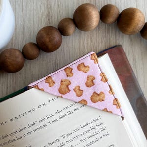 May include: A pink fabric corner bookmark featuring a pattern of brown bear shapes, placed on the pages of a book. The book is open, revealing text. A wooden bead garland and a white ceramic cup are in the background.