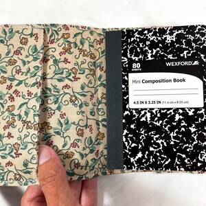 May include: A beige fabric notebook cover with a floral pattern. The cover is open to reveal a black and white composition notebook with 80 sheets. The notebook is labelled "Mini Composition Book" and measures 11.4 cm by 8.25 cm.