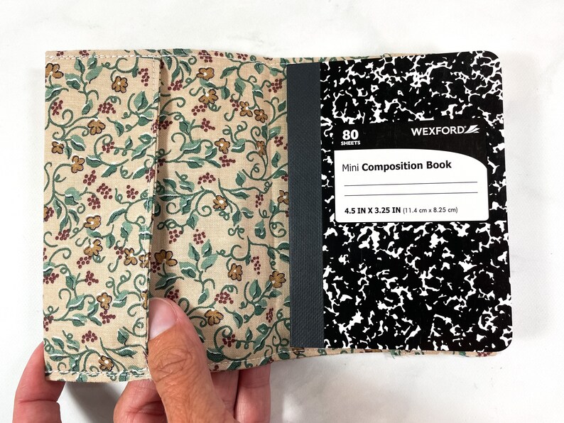 May include: A beige fabric notebook cover with a floral pattern. The cover is open to reveal a black and white composition notebook with 80 sheets. The notebook is labelled "Mini Composition Book" and measures 11.4 cm by 8.25 cm.