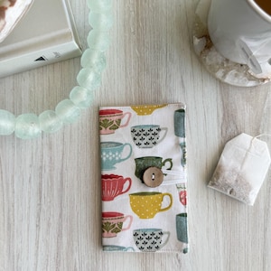 May include: A white fabric pouch with a button closure. The pouch is decorated with a pattern of colorful teacups with floral designs.