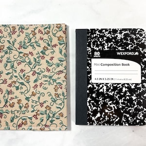 May include: A small composition notebook with a black and white speckled cover. The notebook is 11.4 cm by 8.25 cm and has 80 sheets. The notebook is next to a fabric cover with a floral pattern in shades of brown, green, and red.