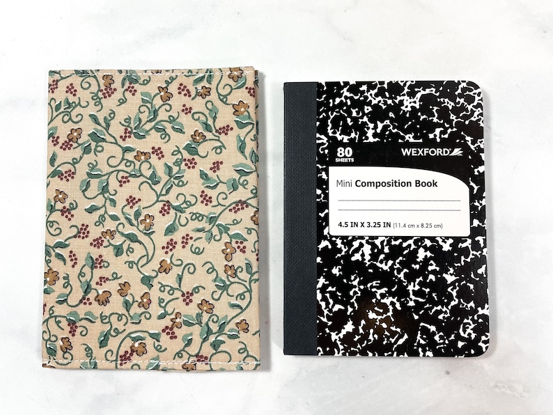 May include: A small composition notebook with a black and white speckled cover. The notebook is 11.4 cm by 8.25 cm and has 80 sheets. The notebook is next to a fabric cover with a floral pattern in shades of brown, green, and red.