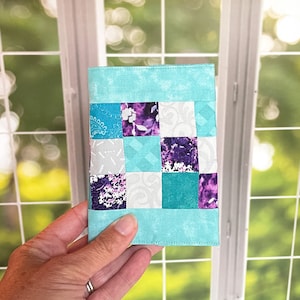 May include: A small, quilted fabric wallet with a patchwork design. The wallet is made of turquoise, white, and purple fabric.
