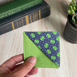 May include: A square fabric bookmark with a green frog pattern on a navy blue background and a solid green corner. The bookmark is held in a hand, with books and a potted plant in the background.