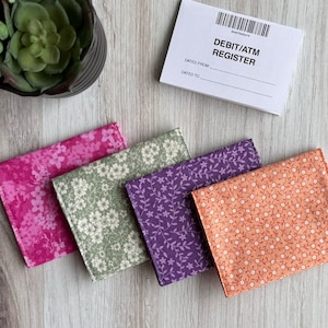 May include: Four fabric pouches with floral patterns. The pouches are pink, green, purple, and orange. The pouches are arranged in a stack, with the pink pouch on top and the orange pouch on the bottom.