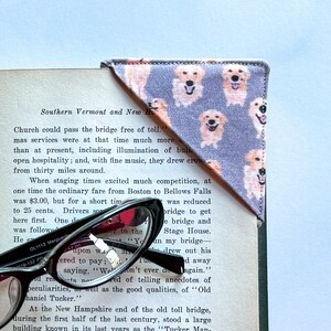 May include: A purple fabric bookmark with a pattern of golden retriever dogs. The bookmark is folded into a triangle shape and is placed on a book with a pair of glasses on top of the book.
