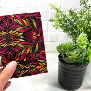 May include: A rectangular fabric item with a vibrant floral pattern in shades of red, orange, and gold on a black background. The design features stylized leaves and a symmetrical arrangement. The item is held in a hand.