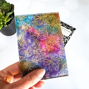 May include: A colorful fabric notebook cover with a gold butterfly design. The cover has a watercolor effect with shades of purple, blue, green, and pink.