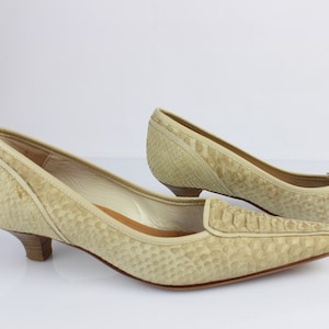 Op de afbeelding: Een paar lichtbeige pumps met een textuur die op reptielen lijkt. De schoenen hebben een spitse neus, een lage hak en een instapontwerp. De hakken zijn ongeveer 3,8 cm hoog.