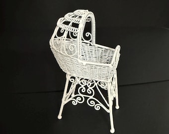 May include: A white wicker cradle featuring an arched hood and a woven basket body. The structure is supported by four slender legs connected by decorative scrollwork detailing, creating a raised stand for the main basket compartment.