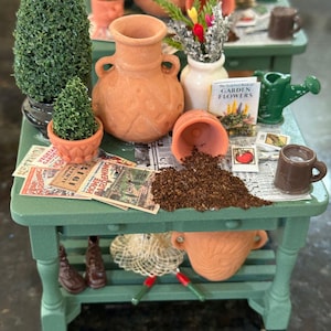 May include: Miniature garden scene with a green table, terracotta pots, and small plants. A small book titled "Garden Flowers" is visible, along with a watering can and a tiny mug. The scene evokes a charming, handcrafted aesthetic.