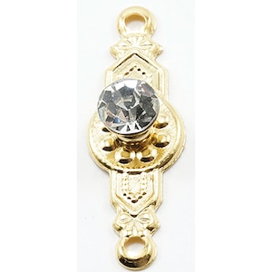 May include: A gold-colored metal charm with a clear, faceted gemstone in the center. The charm has an ornate design with decorative details and loops at the top and bottom for attachment. The charm is likely used for jewelry making or crafting.
