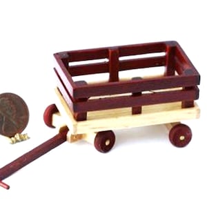 May include: A miniature wooden wagon with a red-brown body and natural wood base. The wagon has four small wheels and a long handle. A penny is next to the wagon for scale. The wagon is likely a decorative item or toy.
