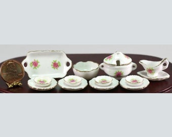 May include: Miniature white porcelain dollhouse dinnerware set with pink rose floral design. Includes a serving tray, bowls, plates, a lidded soup tureen, and a gravy boat. Gold trim accents the edges of each piece. A penny is included for scale.