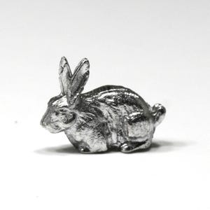 May include: A small, silver-colored rabbit figurine. The bunny is in a seated position with its ears upright. The figurine has detailed features and a shiny finish, set against a plain white background.