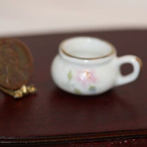 May include: A miniature white porcelain teacup with a handle and gold trim, decorated with pink and blue floral accents. A penny is next to the teacup for scale. The teacup is on a dark brown surface. This item is likely a dollhouse accessory.