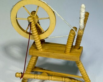 May include: A miniature wooden spinning wheel featuring a light tan finish, a circular wheel with spokes, a vertical spindle topped with white fiber, and a thin red wire connecting the wheel to the base, all supported by turned wooden legs.