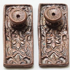 May include: Two antique bronze-colored cabinet knobs. Each knob has a rectangular base with an ornate, symmetrical design and a circular knob on top. The knobs are likely made of metal and show signs of age and wear.