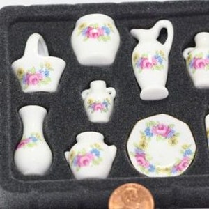 May include: A set of miniature white porcelain dishes with floral designs in pink, blue, and yellow. The set includes a basket, vases, a pitcher, a plate, and a bottle, all arranged in a black tray.