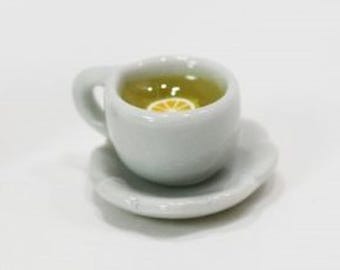 May include: A miniature white ceramic teacup and saucer set containing a translucent yellow liquid with a small, circular yellow and white lemon slice floating on the surface. The cup features a curved handle and a smooth, glossy finish.