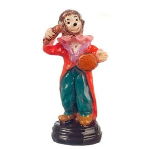 May include: A small figurine of a monkey dressed in a red coat, teal trousers, and a pink ruffled collar. The monkey holds a brown object in one hand and a round orange item in the other, standing on a dark brown circular base.
