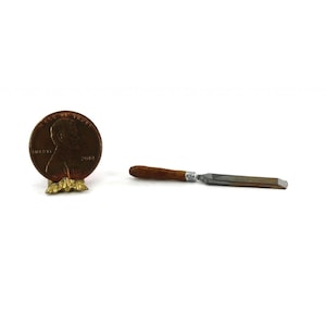 May include: A miniature chisel with a wooden handle and a metal blade, next to a penny. The chisel is a tool used for woodworking or carving. The penny is a U.S. one-cent coin.