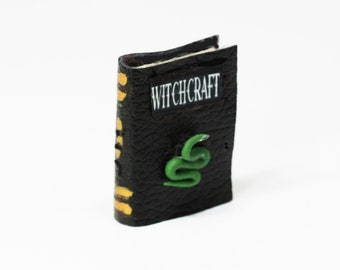 May include: A miniature black book with the word "WITCHCRAFT" in white lettering. A green snake is on the cover. The spine has gold accents. The book is likely a decorative item or part of a dollhouse collection.