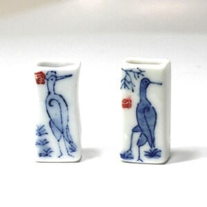 May include: Two small, rectangular white ceramic vases. Each vase features a blue illustration of a bird with a long beak and a red stamp. The bird is standing on a patch of grass. The vases are likely for decorative purposes.